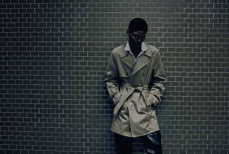 A Black man leans against a gray tiled wall wearing a tan trench coat and dark pants in a medium shot with neutral lighting.