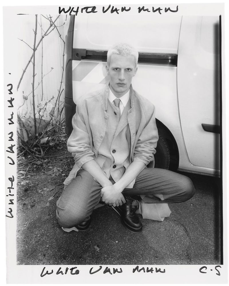 Monochrome portrait of a crouched adult male with fair hair wearing a blazer and dress pants, posing near a white van and bare tree branches.