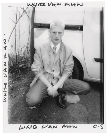Monochrome portrait of a crouched adult male with fair hair wearing a blazer and dress pants, posing near a white van and bare tree branches.