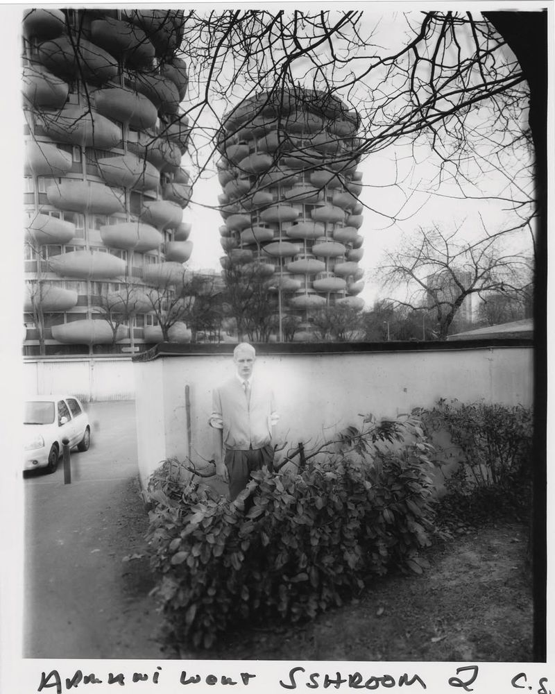 Black and white photo of a man in front of a wall and bushes, with two rounded apartment buildings visible in the background.