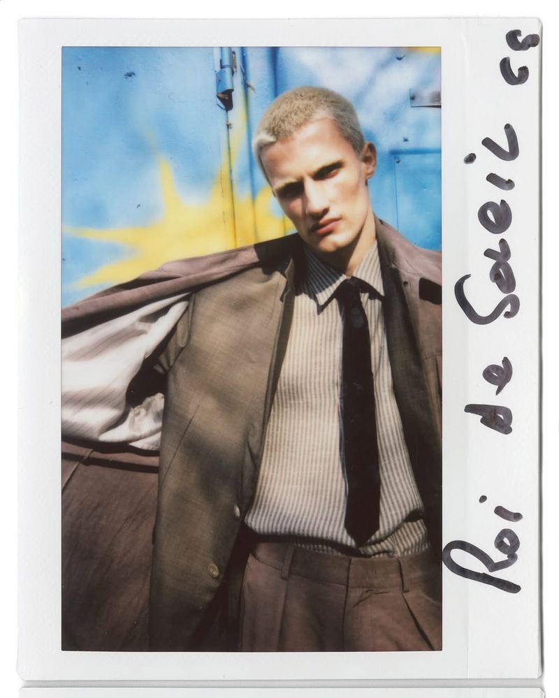 Man with bleached hair and tan suit jacket poses in front of a blue and yellow painted background; fashion portrait.