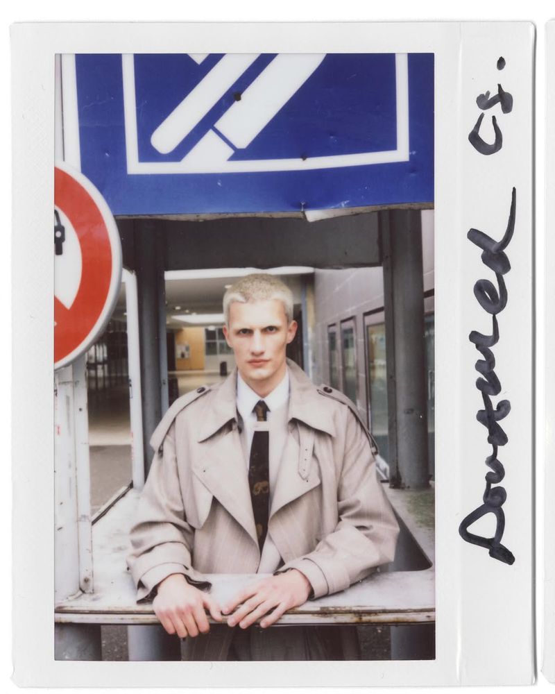 Studio-style portrait of a young adult male with light hair, wearing a tan overcoat and tie, standing indoors near signage.