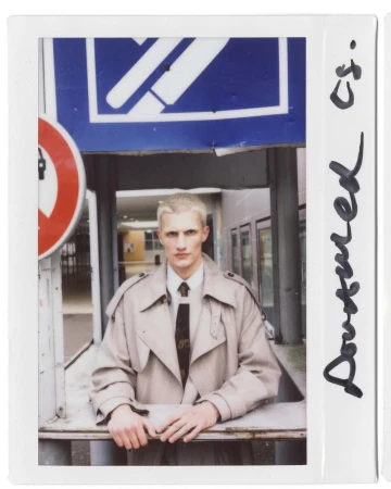 Studio-style portrait of a young adult male with light hair, wearing a tan overcoat and tie, standing indoors near signage.