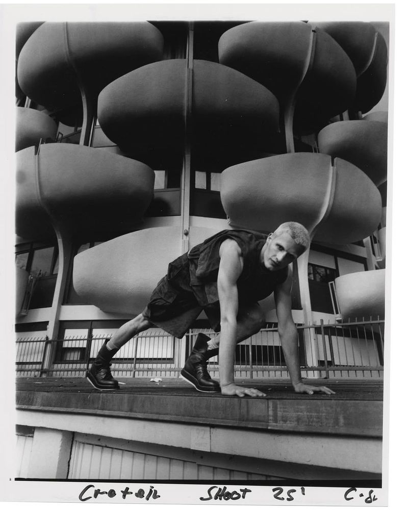 Model crouching outdoors in front of a modern building with rounded balconies wearing dark clothing and boots