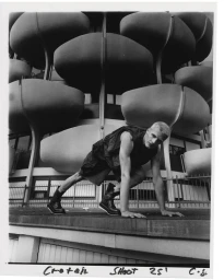 Model crouching outdoors in front of a modern building with rounded balconies wearing dark clothing and boots