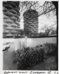 Black and white editorial portrait of a male model standing outdoors in front of tall rounded residential buildings.