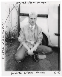 Monochrome portrait of a male model crouching outdoors next to a white van wearing a suit jacket and tie