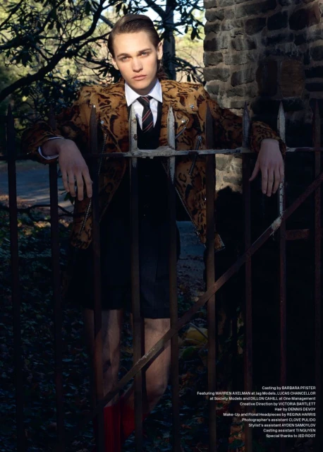 Portrait of a young adult male in an patterned jacket, tie, and red boots leaning on a metal gate in an outdoor setting.
