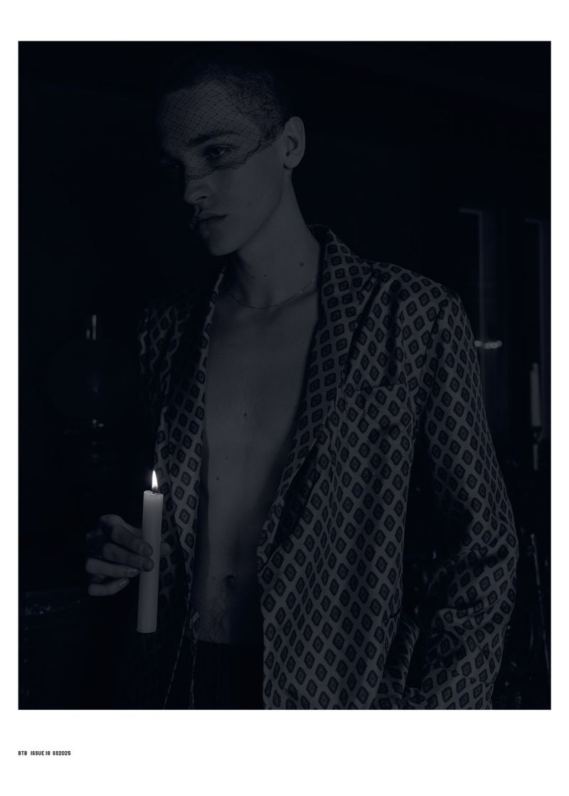 Portrait of a young adult male in patterned jacket holding lit candle in a dark room. He has a sheer veil over his face.