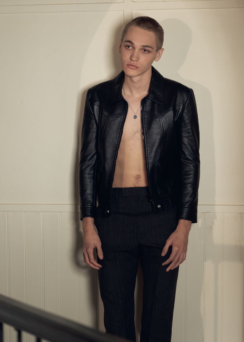 Portrait of a young adult man wearing a black leather jacket with no shirt, black pants, and silver chain posing indoors.