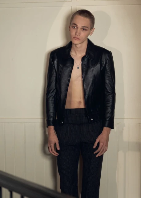 Portrait of a young adult man wearing a black leather jacket with no shirt, black pants, and silver chain posing indoors.