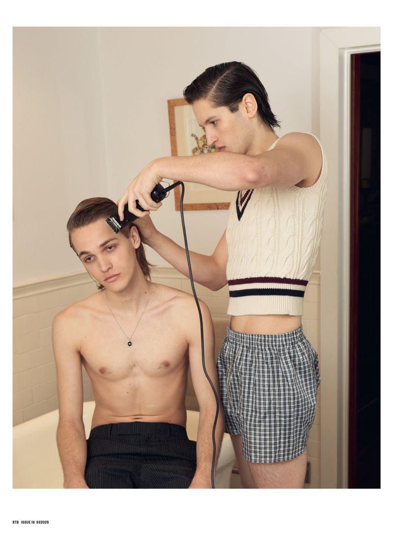 Two young adult males in a bathroom: one giving the other a haircut using electric clippers, lit by indoor lighting.
