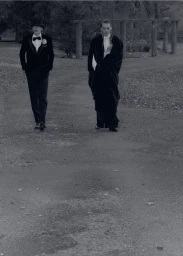 Two men in formal evening wear walking outdoors along a gravel path in a park for Behind the Blinds editorial