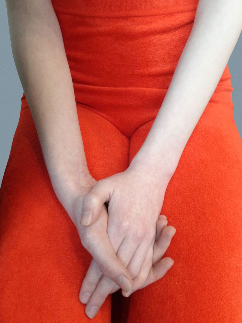 Close-up studio shot of a fair-skinned adult with crossed hands in front of their red-orange outfit and a simple grey background.