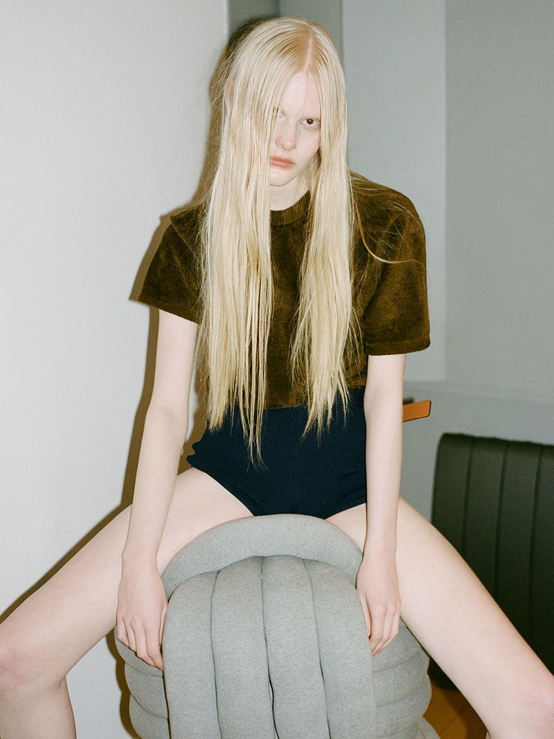 Blonde woman wearing a brown shirt and black underwear sitting on a gray ottoman in an indoor photo studio.