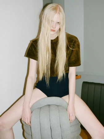 Blonde woman wearing a brown shirt and black underwear sitting on a gray ottoman in an indoor photo studio.