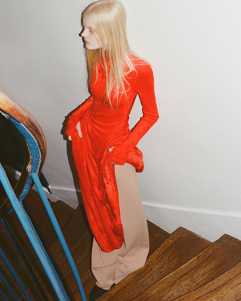 A fair-skinned woman with long blonde hair descends a staircase in a red velvet dress and tan pants in a low-angle, indoor fashion shot.