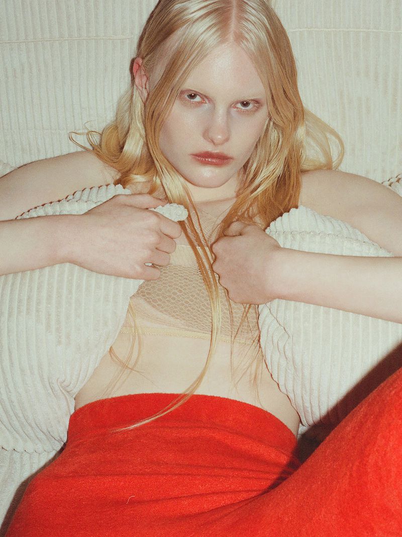 A fair-skinned young adult female with blonde hair reclines against a white corduroy surface, wearing a beige top and red trousers.