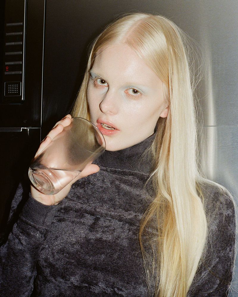Close-up of a woman with platinum blonde hair drinking water from a glass against a muted background. Beauty and hydration concept.