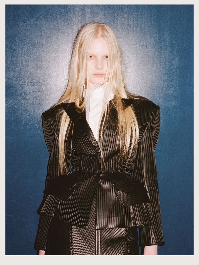 Fashionable woman with long blonde hair poses in a structured black blazer and skirt against a textured blue wall, creating a striking contrast.