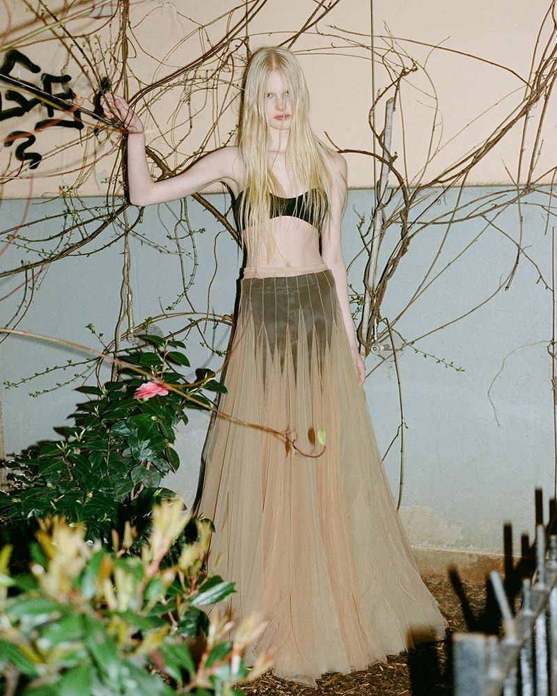 Model with long blonde hair wearing a black crop top and sheer beige tulle skirt standing outdoors near bare branches