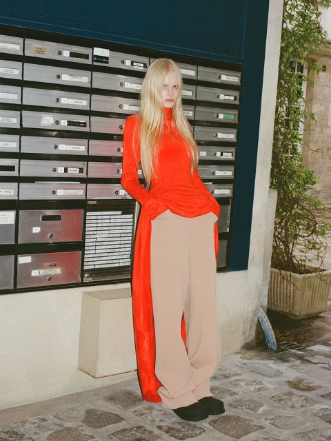 A woman with long blonde hair, wearing a bright red turtleneck and beige pants, poses in front of an apartment building with mailboxes.