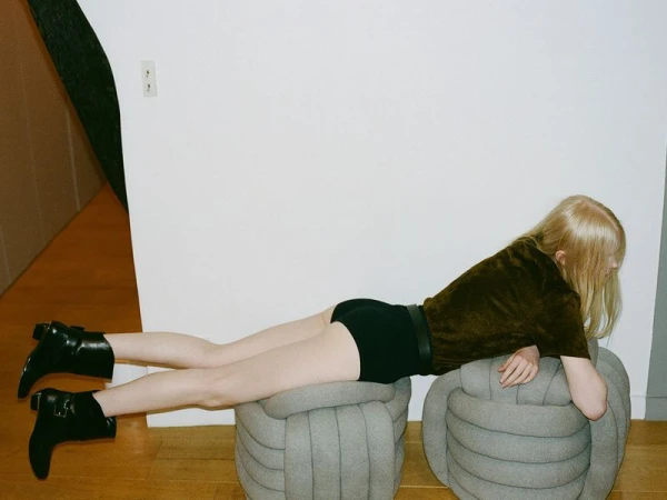An adult woman with blonde hair poses on a light grey ottoman wearing black shorts, a brown top, and black boots in a simple indoor space.