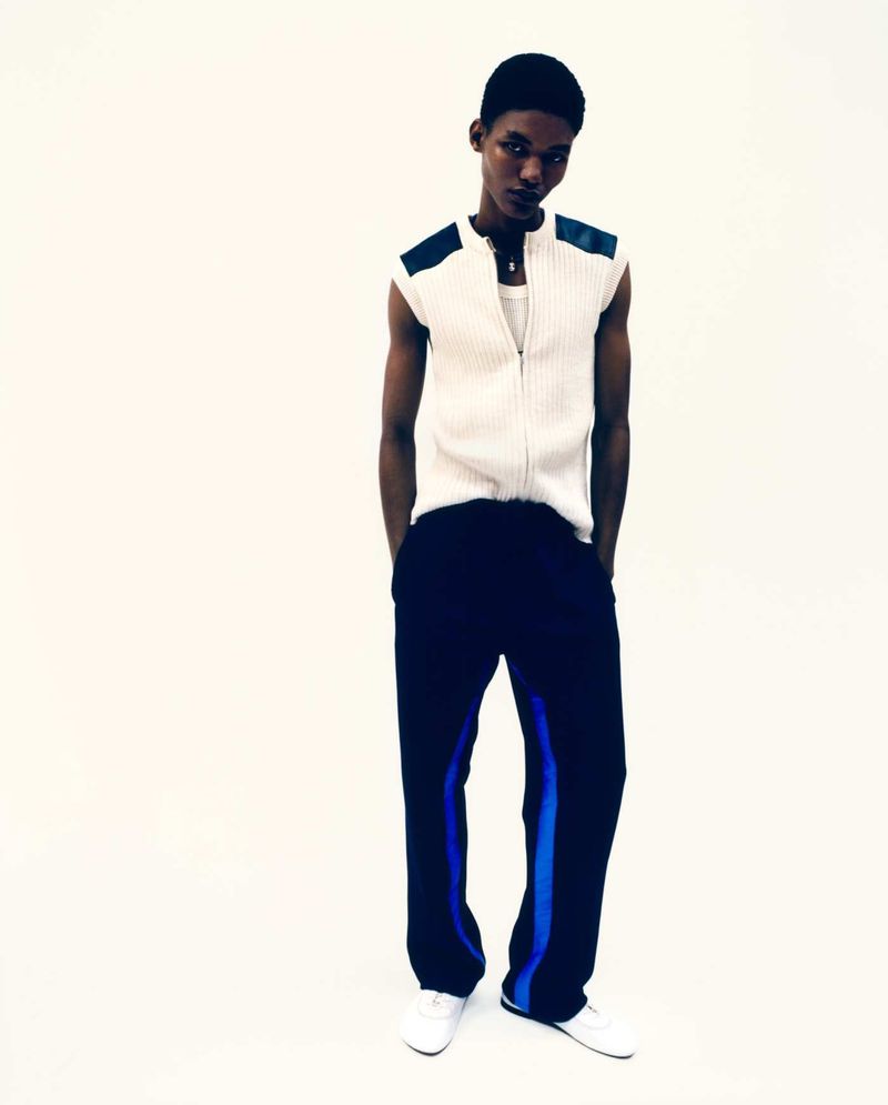 A young Black man wearing a white ribbed vest and navy blue track pants stands against a white studio background.
