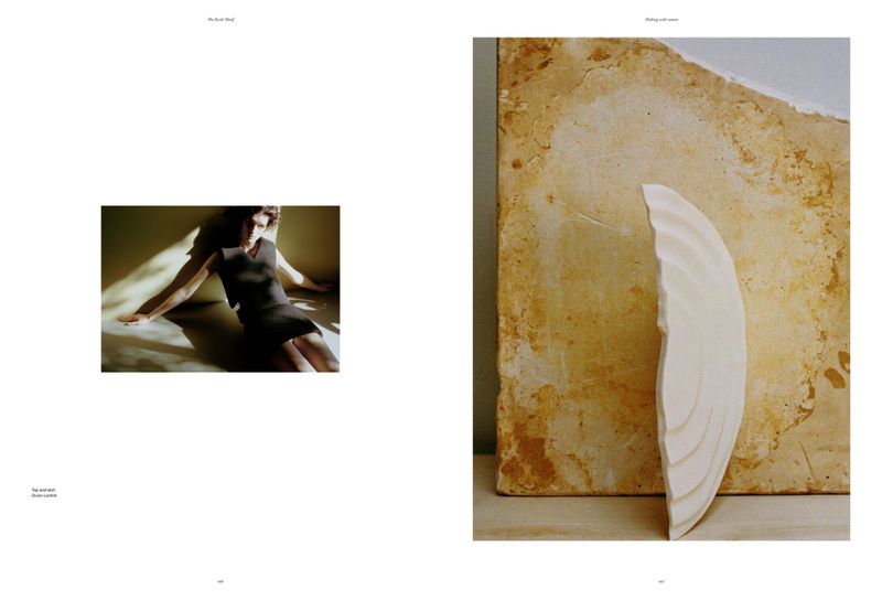 Woman in a dark dress poses in a sunlit corner in a fashion editorial shot, next to a textural plaster wall fragment sculpture.