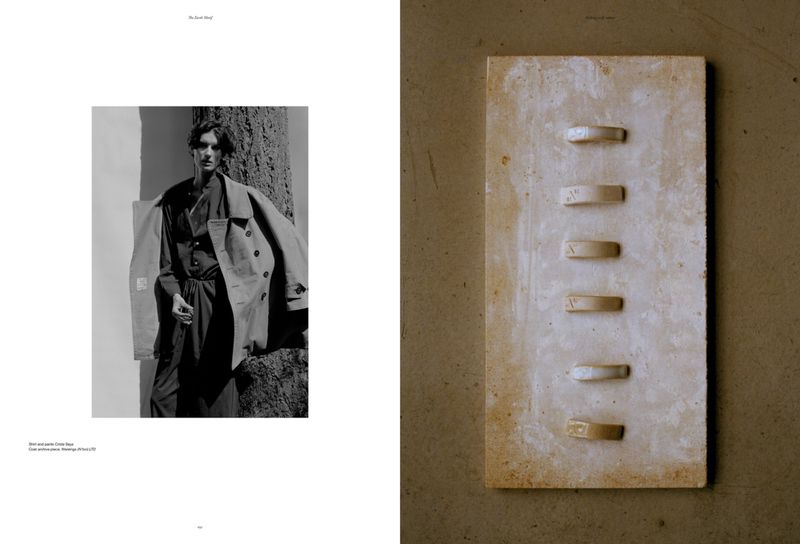 Black and white fashion shot of a man in a suit with an overcoat next to a tree, and a light beige textured stone plaque.
