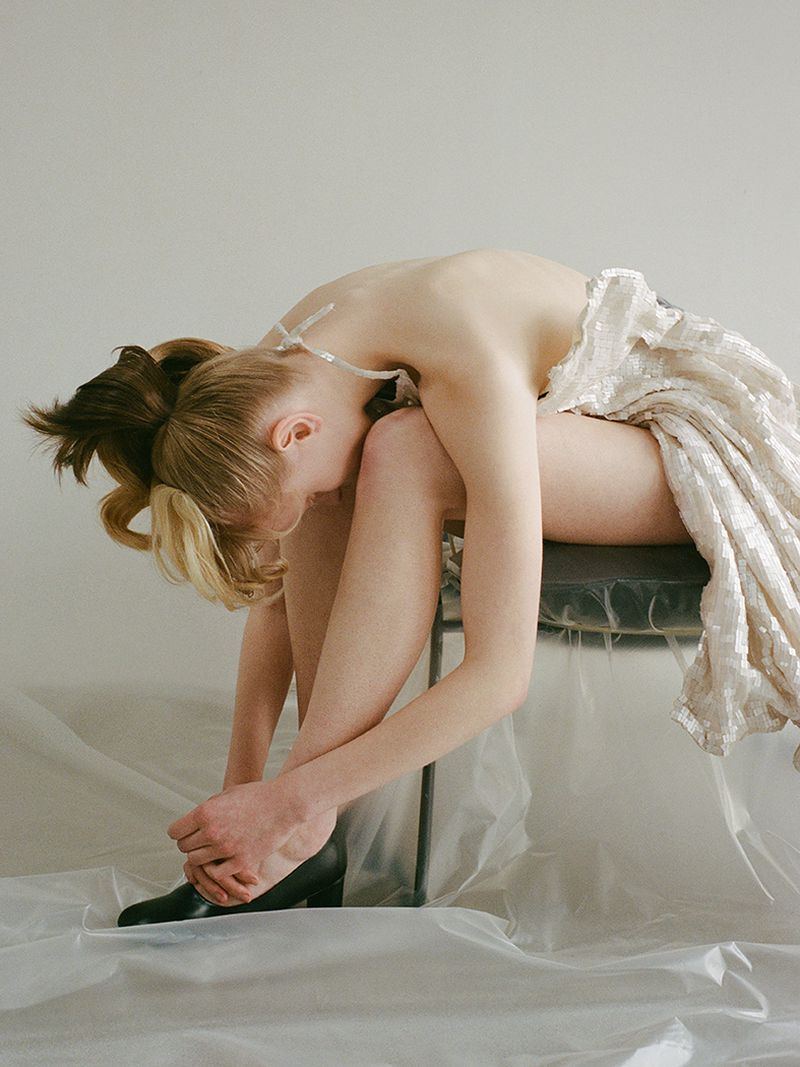 Young woman with blonde and brunette hair sits hunched over on a stool wearing a light colored dress and dark shoes in a minimalist studio.