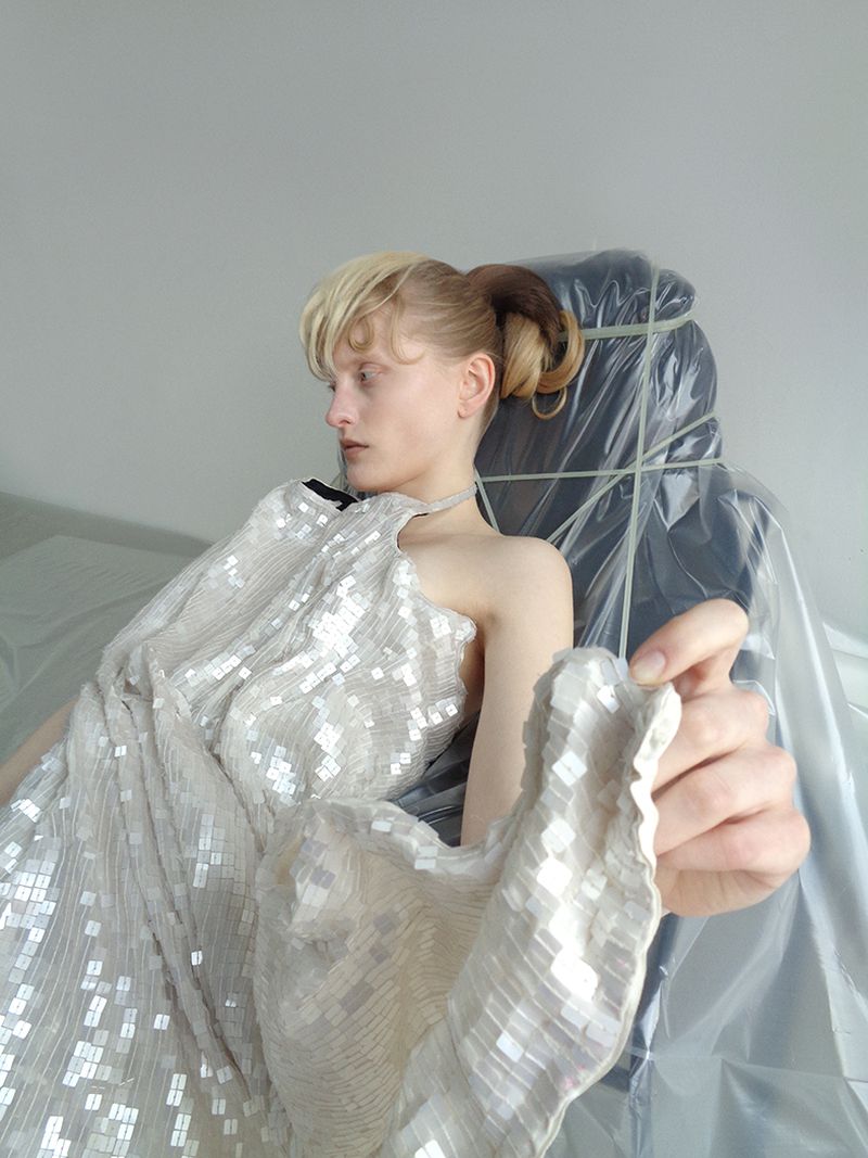 Model in a sequined gown poses near an object wrapped in plastic. Studio lighting highlights gown's texture, indoor fashion shot.