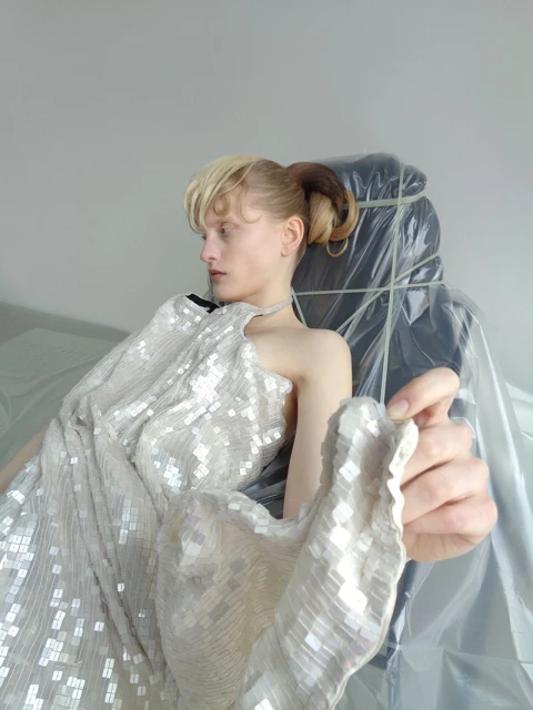 Model in a sequined gown poses near an object wrapped in plastic. Studio lighting highlights gown's texture, indoor fashion shot.