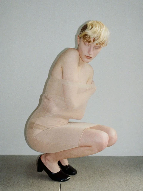 Blonde woman with bare shoulders and a beige dress poses in a squatting position with black high heels in a neutral studio.