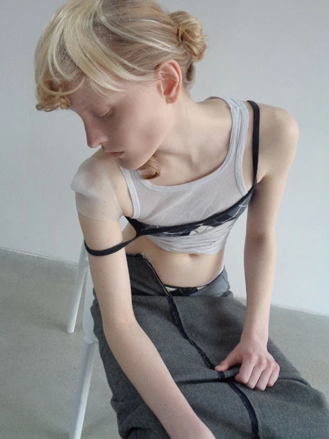 Young woman in a gray skirt and white tank top sits on a chair in front of a gray wall, looking down with a serene expression.