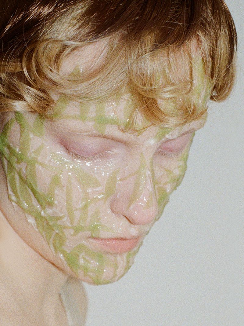 Close-up of a young adult with light skin and blonde hair wearing a green facial mask. The person is looking down, eyes closed.