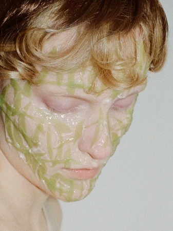 Close-up of a young adult with light skin and blonde hair wearing a green facial mask. The person is looking down, eyes closed.