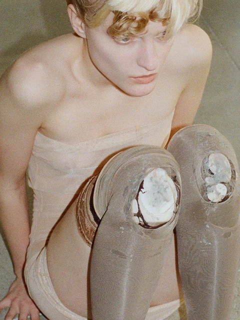 Close-up of a young adult posing in neutral tones, featuring ripped leggings and an unusual textured material, set against a plain backdrop.