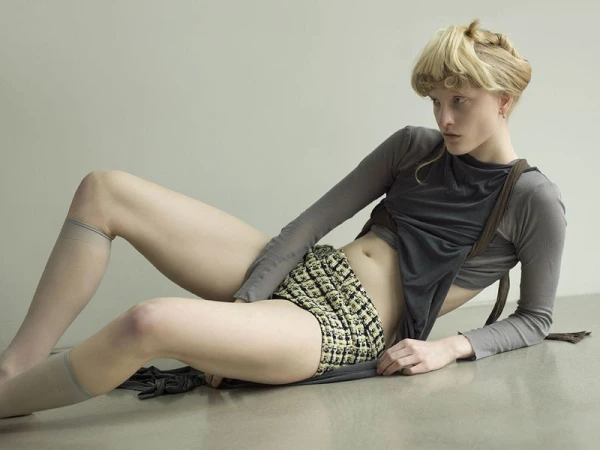 A light-skinned young adult woman with blonde hair poses on the floor in a gray long-sleeved top and patterned shorts, indoors.
