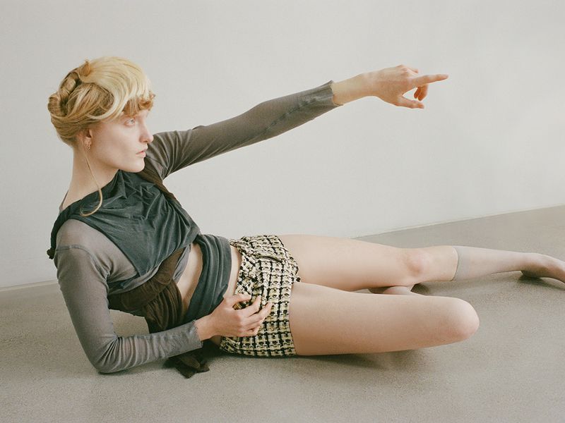 Indoor fashion shot of a young adult female model reclining on a floor, wearing shorts and a top, with her arm raised, pointing.