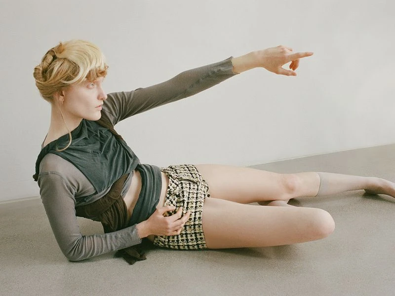 Indoor fashion shot of a young adult female model reclining on a floor, wearing shorts and a top, with her arm raised, pointing.