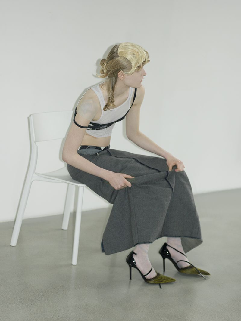 Adult female seated on a chair with blonde hair wearing a white top, grey skirt, and heels in front of a white backdrop in a studio.