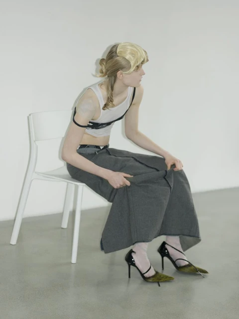 Adult female seated on a chair with blonde hair wearing a white top, grey skirt, and heels in front of a white backdrop in a studio.