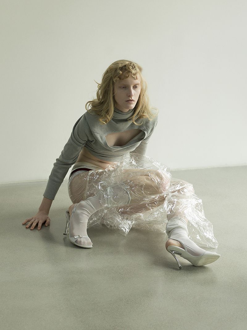 A model poses in a gray crop top and clear plastic wrap, seated against a pale background on a gray floor, wearing white heels.