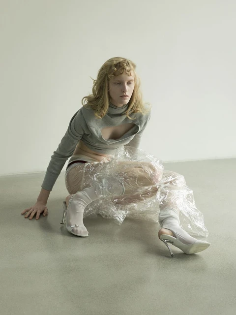 A model poses in a gray crop top and clear plastic wrap, seated against a pale background on a gray floor, wearing white heels.