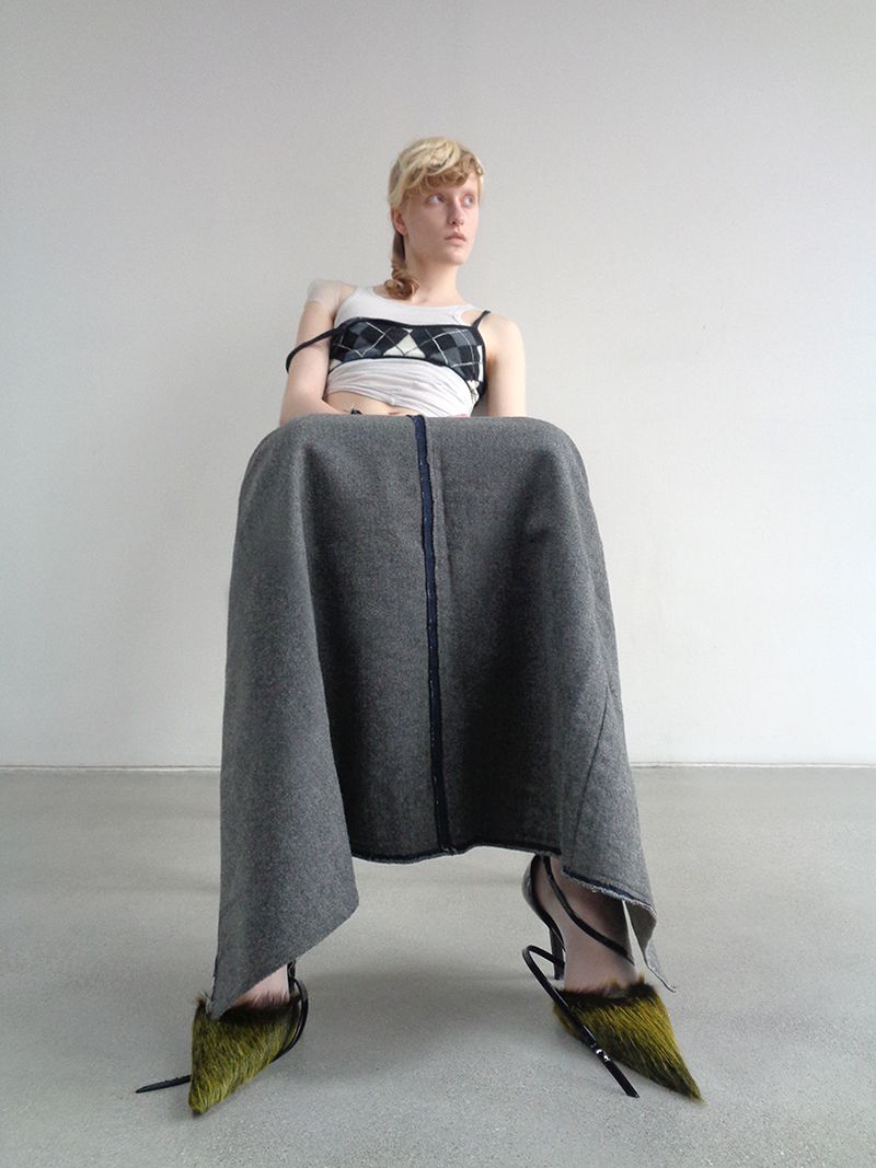 Fashionable woman sitting with a grey skirt, a patterned crop top, and furry high heels against a neutral wall and floor backdrop.