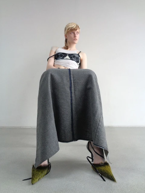 Fashionable woman sitting with a grey skirt, a patterned crop top, and furry high heels against a neutral wall and floor backdrop.