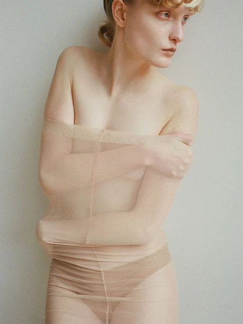 Pale-skinned woman in a nude mesh garment strikes a contemplative pose indoors against a pale wall, soft lighting, and a simple hairstyle.