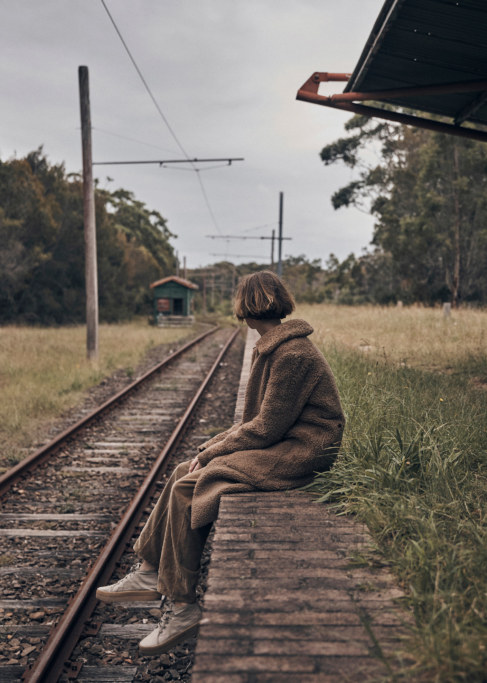 Woman sits on the edge of a train platform wearing a brown coat and pants with the railway tracks leading to a small green building.