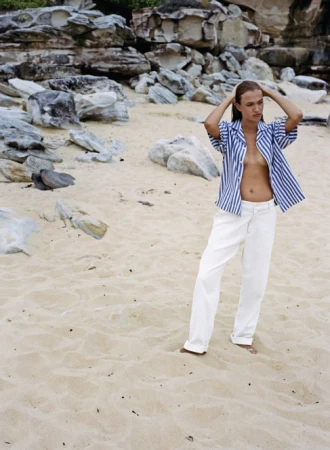 Model standing on a sandy beach wearing a blue and white striped shirt and white trousers with rocky outcrops behind.